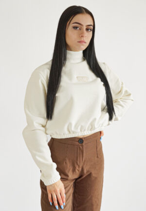A model wearing a cream-colored high-neck crop sweatshirt with a mock neck, elasticated waist, and a subtle "NEAT" logo embroidery on the chest, paired with brown trousers.