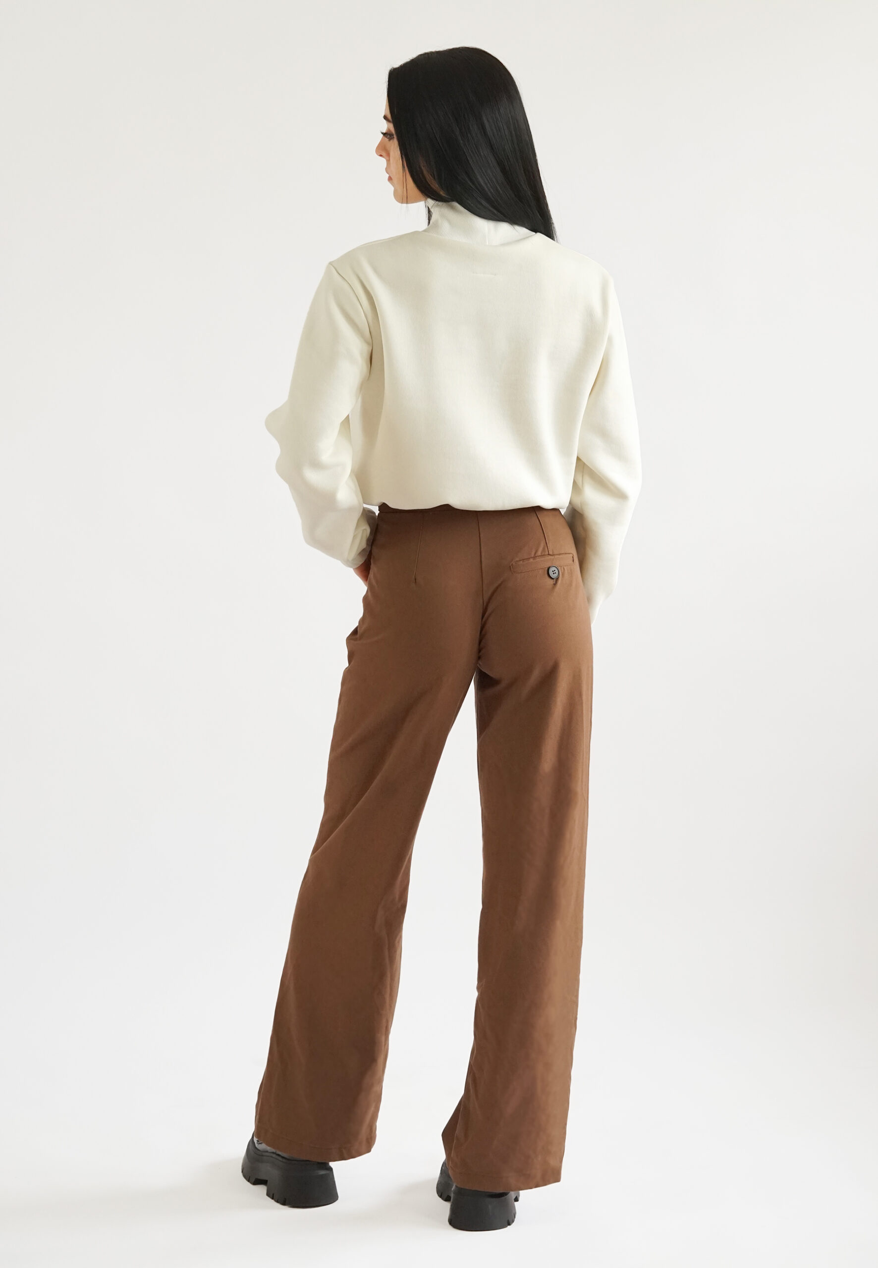 back view A model wearing a cream-colored high-neck crop sweatshirt with a mock neck, elasticated waist, and a subtle "NEAT" logo embroidery on the chest, paired with brown trousers.