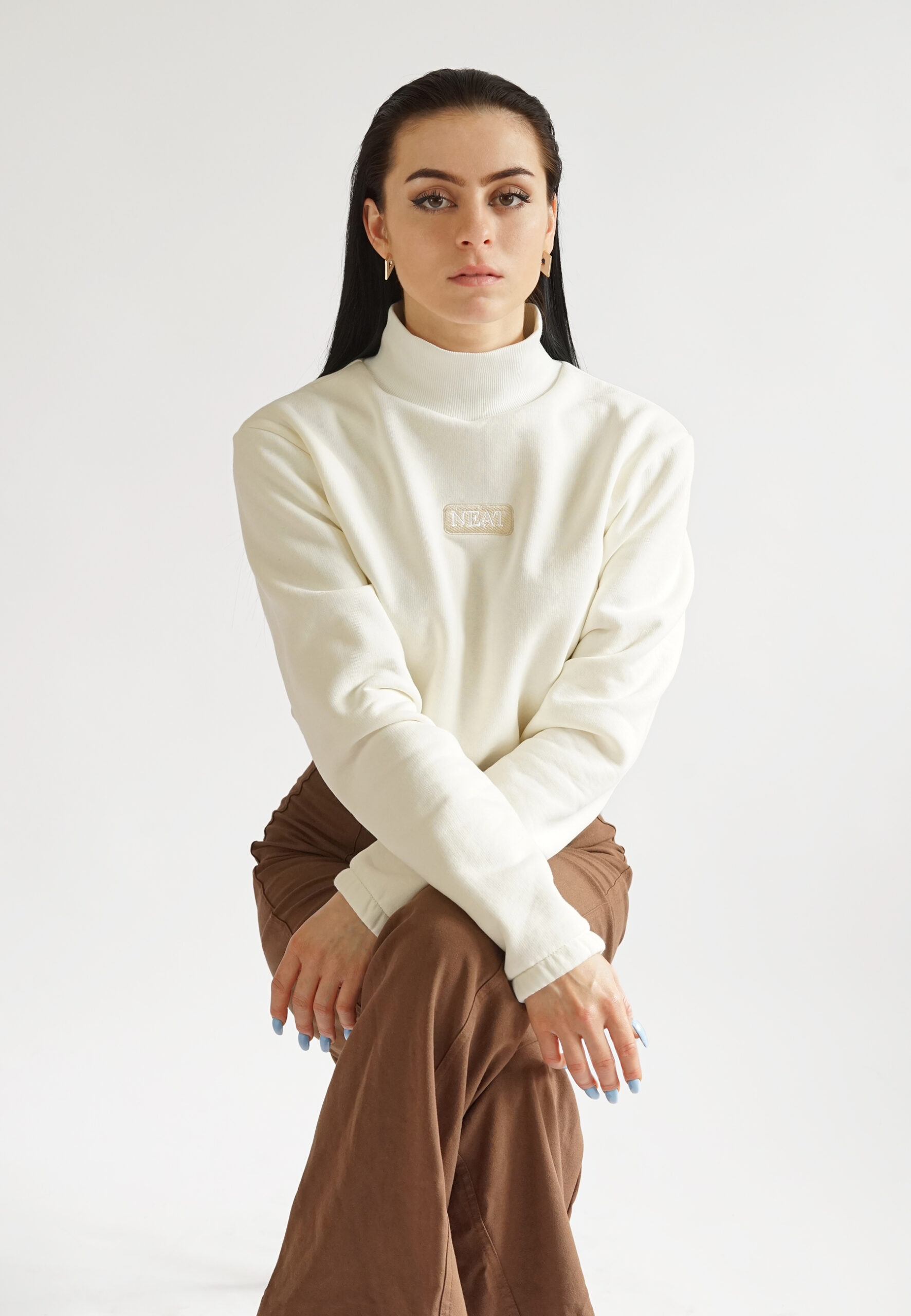 A studio portrait of a model sitting and posing in a cream high-neck crop sweatshirt with a mock collar and "NEAT" logo embroidery, styled with brown high-waisted trousers.celebrity inspired look