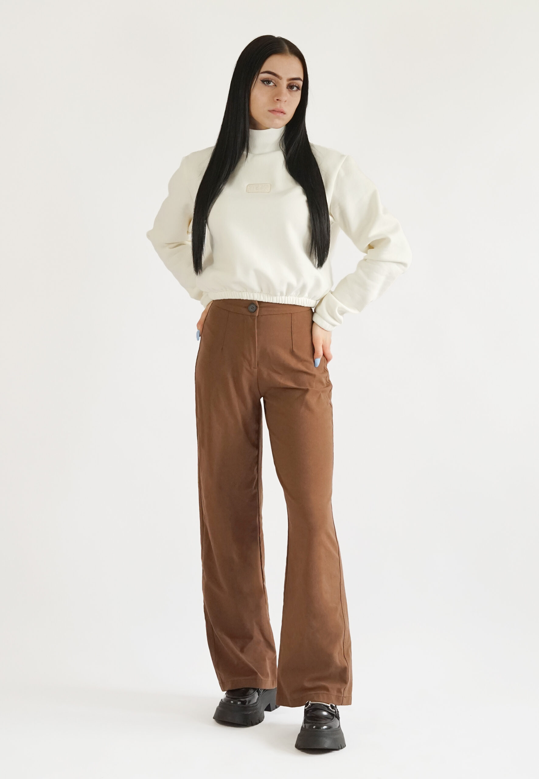 front view A model wearing a cream-colored high-neck crop sweatshirt with a mock neck, elasticated waist, and a subtle "NEAT" logo embroidery on the chest, paired with brown trousers.