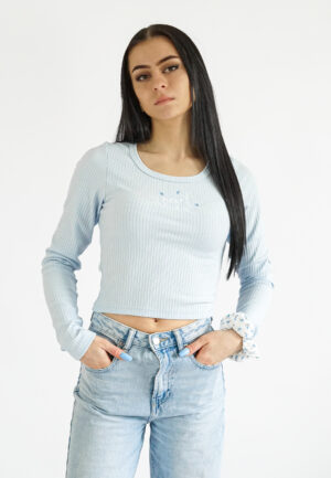 Model wearing a light blue, long-sleeved ribbed crop top with a small central embroidery, paired with light wash denim jeans.