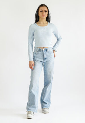 Front view of a model wearing a light blue long-sleeved ribbed crop top with a small embroidery on the chest, paired with light blue denim jeans.