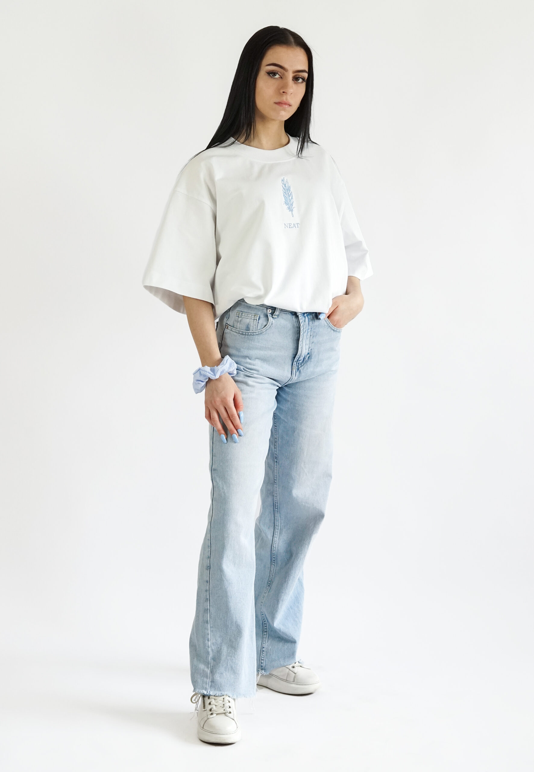 A full-body studio shot of a model wearing a complete casual outfit featuring a white oversized t-shirt with blue embroidery, light wash denim jeans, and a matching blue scrunchie.