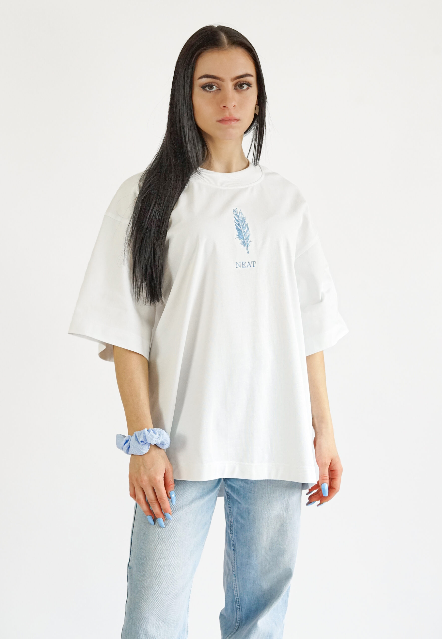Front view of a model wearing a white oversized t-shirt featuring a delicate light blue feather embroidery and the "NEAT" brand name on the chest.