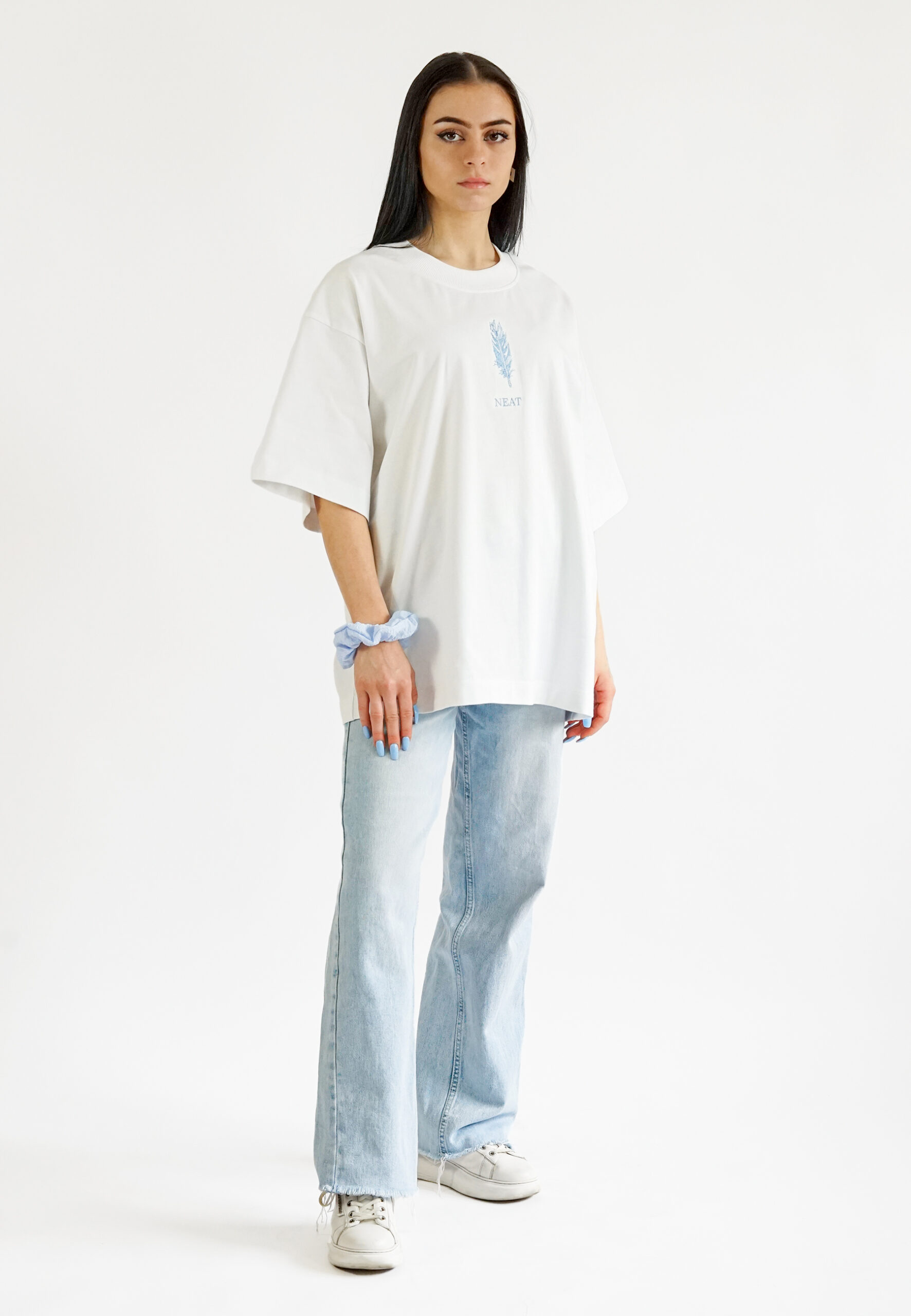 Full-length front view of a model wearing a white oversized t-shirt with a blue feather embroidery, paired with light blue denim jeans.