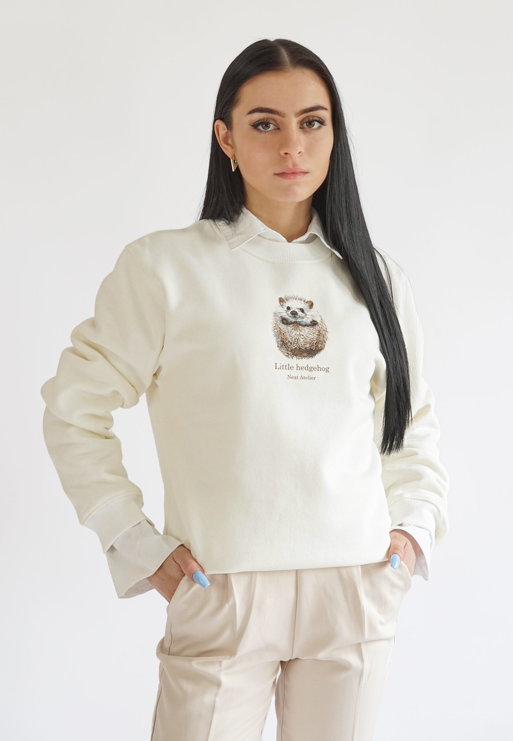 A model wearing a cream-colored crewneck sweatshirt featuring a cute "Little hedgehog" illustration and Neat Atelier branding on the chest, layered over a white collared shirt.