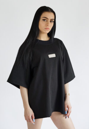 A model wearing a black oversized t-shirt with dropped shoulders and a minimalist rectangular NEAT logo embroidered on the chest, styled as a streetwear piece.