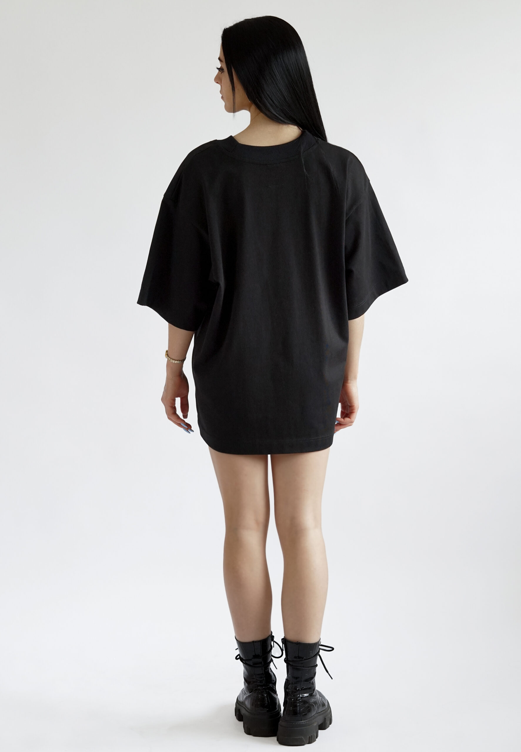 photo from behind A model wearing a black oversized t-shirt with dropped shoulders and a minimalist rectangular NEAT logo embroidered on the chest, styled as a streetwear piece.