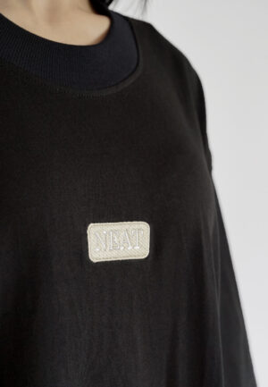 Detail black oversized t-shirt with dropped shoulders and a minimalist rectangular NEAT logo embroidered on the chest, styled as a streetwear piece.