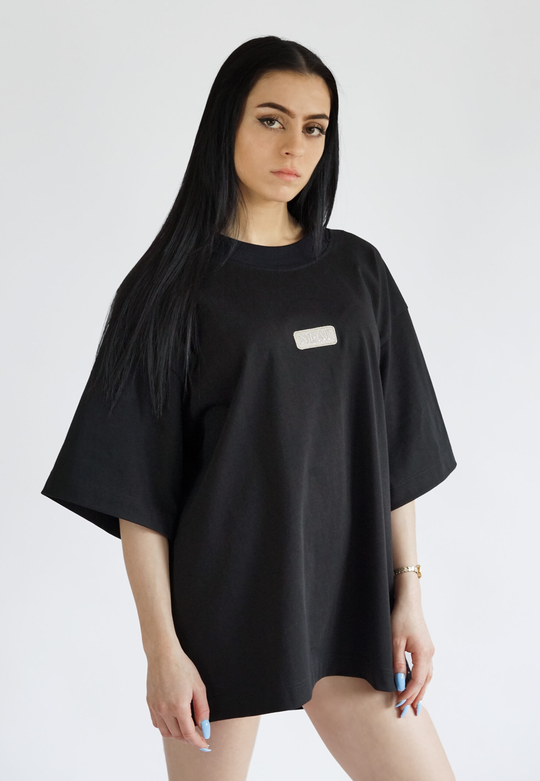 A model wearing a black oversized t-shirt with dropped shoulders and a minimalist rectangular NEAT logo embroidered on the chest, styled as a streetwear piece.