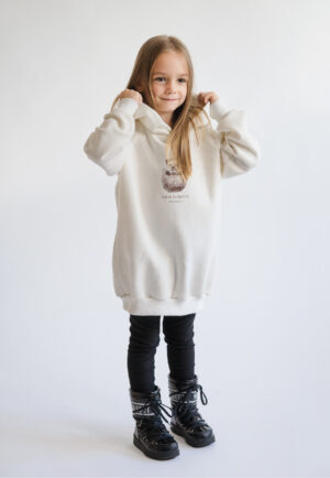 A young girl smiling and wearing a cream-colored oversized hoodie with a "Little hedgehog" illustration and Neat Atelier branding, designed for a matching mother-and-daughter look. moon boots