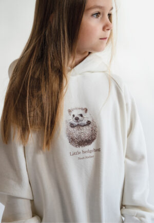 A young girl smiling and wearing a cream-colored oversized hoodie with a "Little hedgehog" illustration and Neat Atelier branding, designed for a matching mother-and-daughter look. detail moon boots