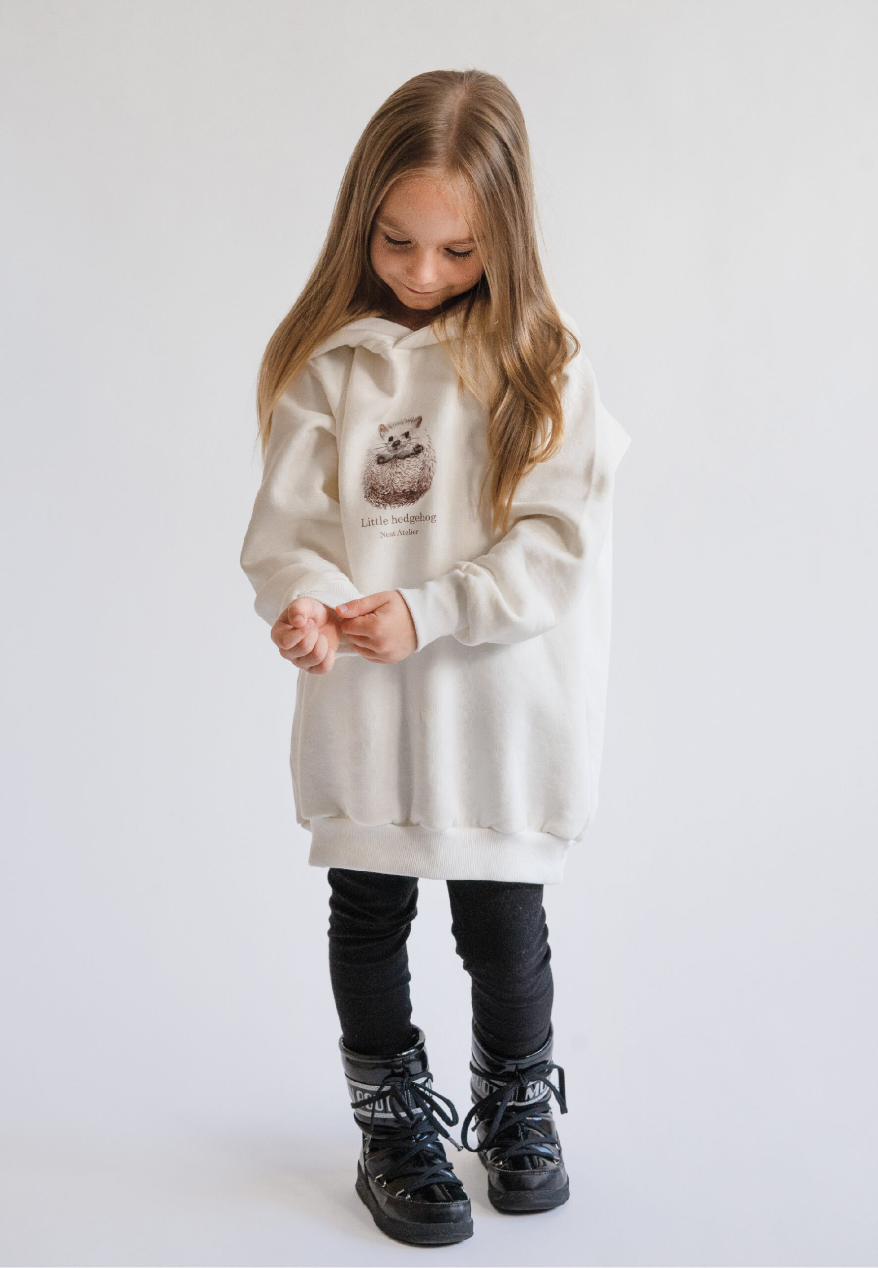 A young girl smiling and wearing a cream-colored oversized hoodie with a "Little hedgehog" illustration and Neat Atelier branding, designed for a matching mother-and-daughter look. outfit moon boots