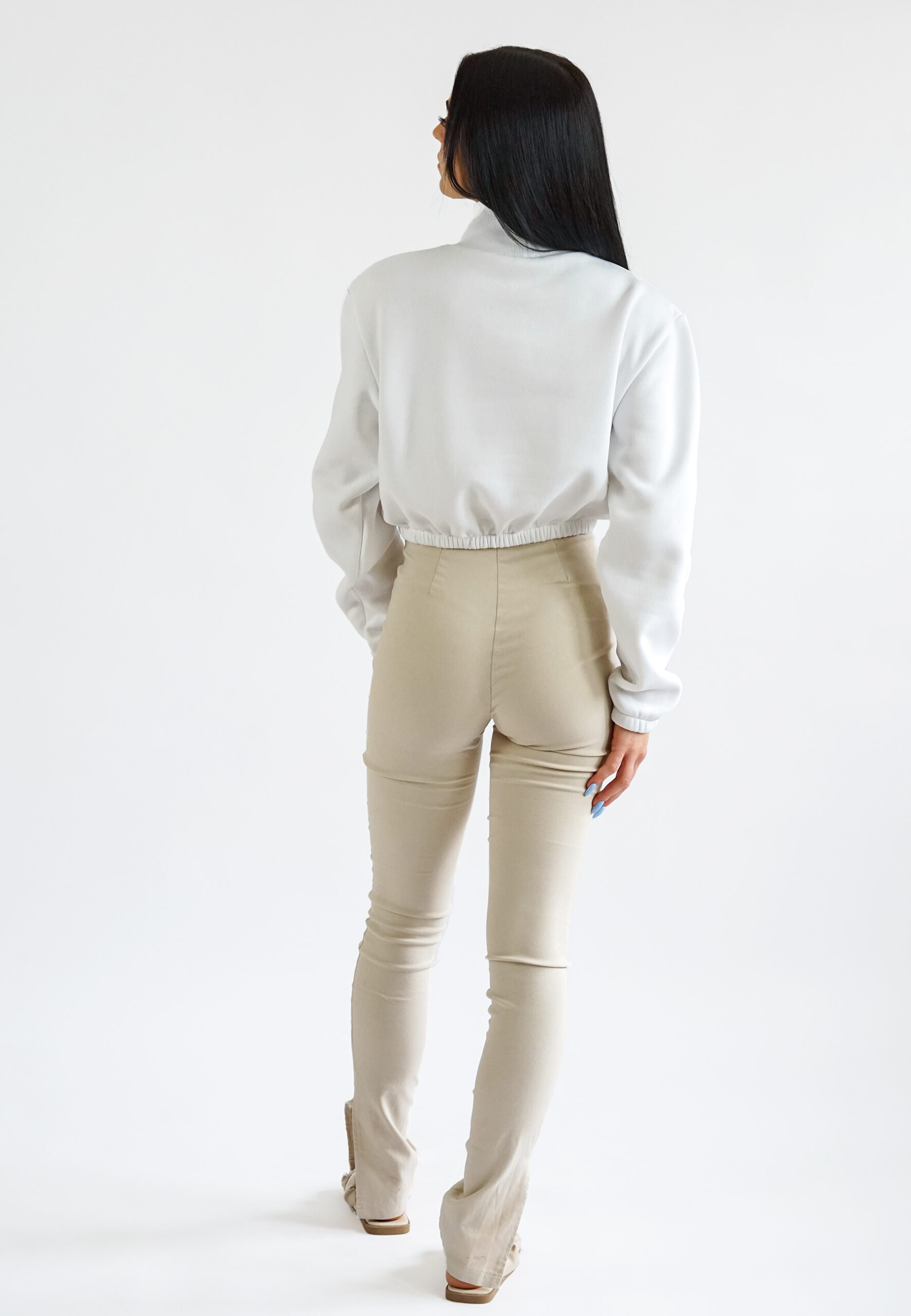 A model wearing a white high-neck crop sweatshirt featuring a vibrant blue floral print on the chest, paired with beige high-waisted trousers.