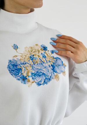 A model wearing a white high-neck crop sweatshirt featuring a vibrant blue floral print on the chest, paired with beige high-waisted trousers. detail