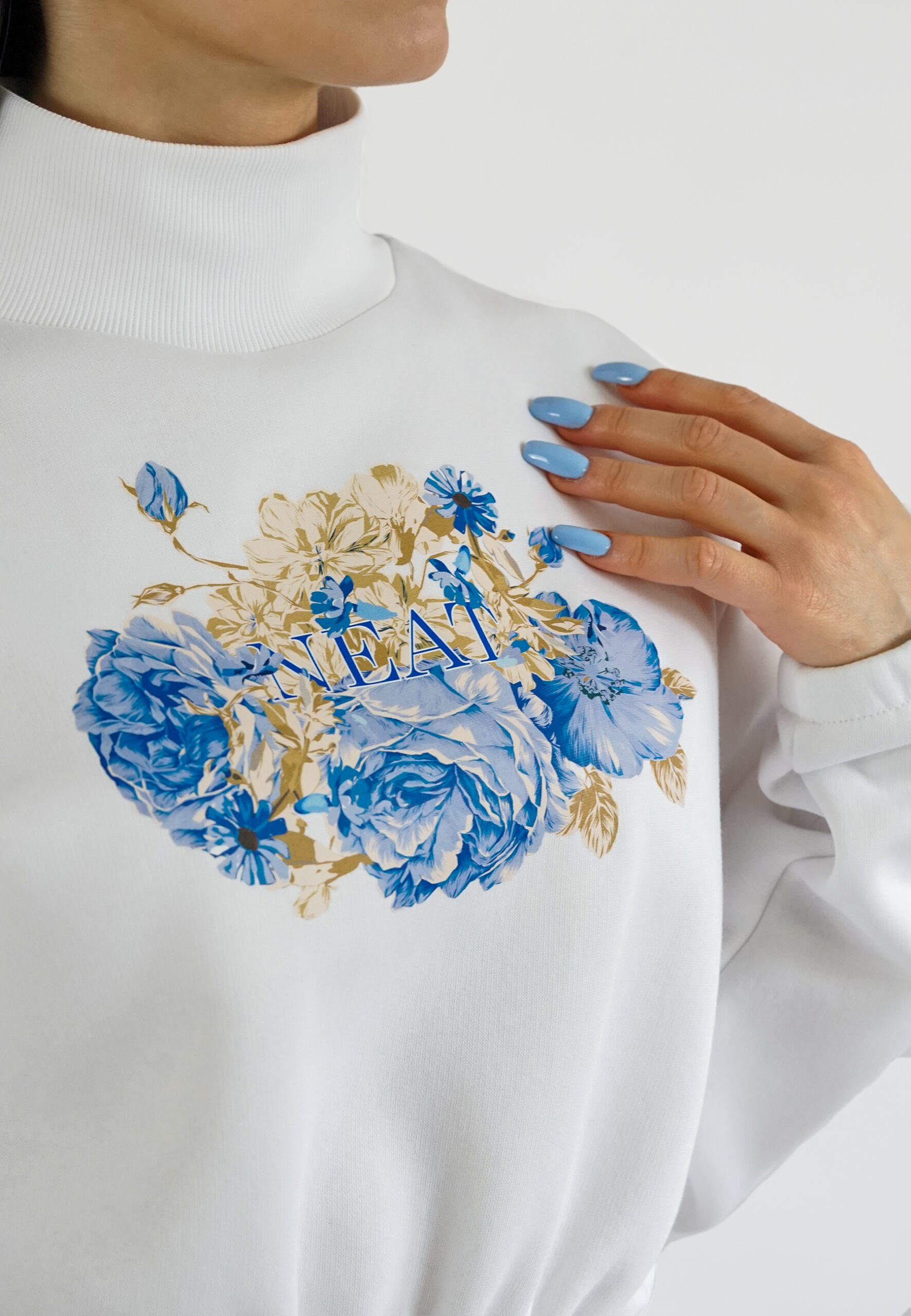 A model wearing a white high-neck crop sweatshirt featuring a vibrant blue floral print on the chest, paired with beige high-waisted trousers. detail