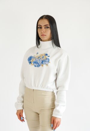 A model wearing a white high-neck crop sweatshirt featuring a vibrant blue floral print on the chest, paired with beige high-waisted trousers.