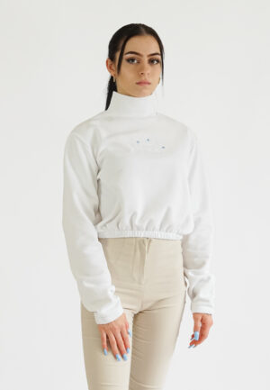 A model wearing a white high-neck crop sweatshirt with a delicate blue floral embroidery and NEAT logo on the chest, paired with beige trousers.