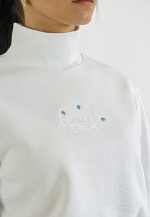 Detail white high-neck crop sweatshirt with a delicate blue floral embroidery and NEAT logo on the chest, paired with beige trousers.