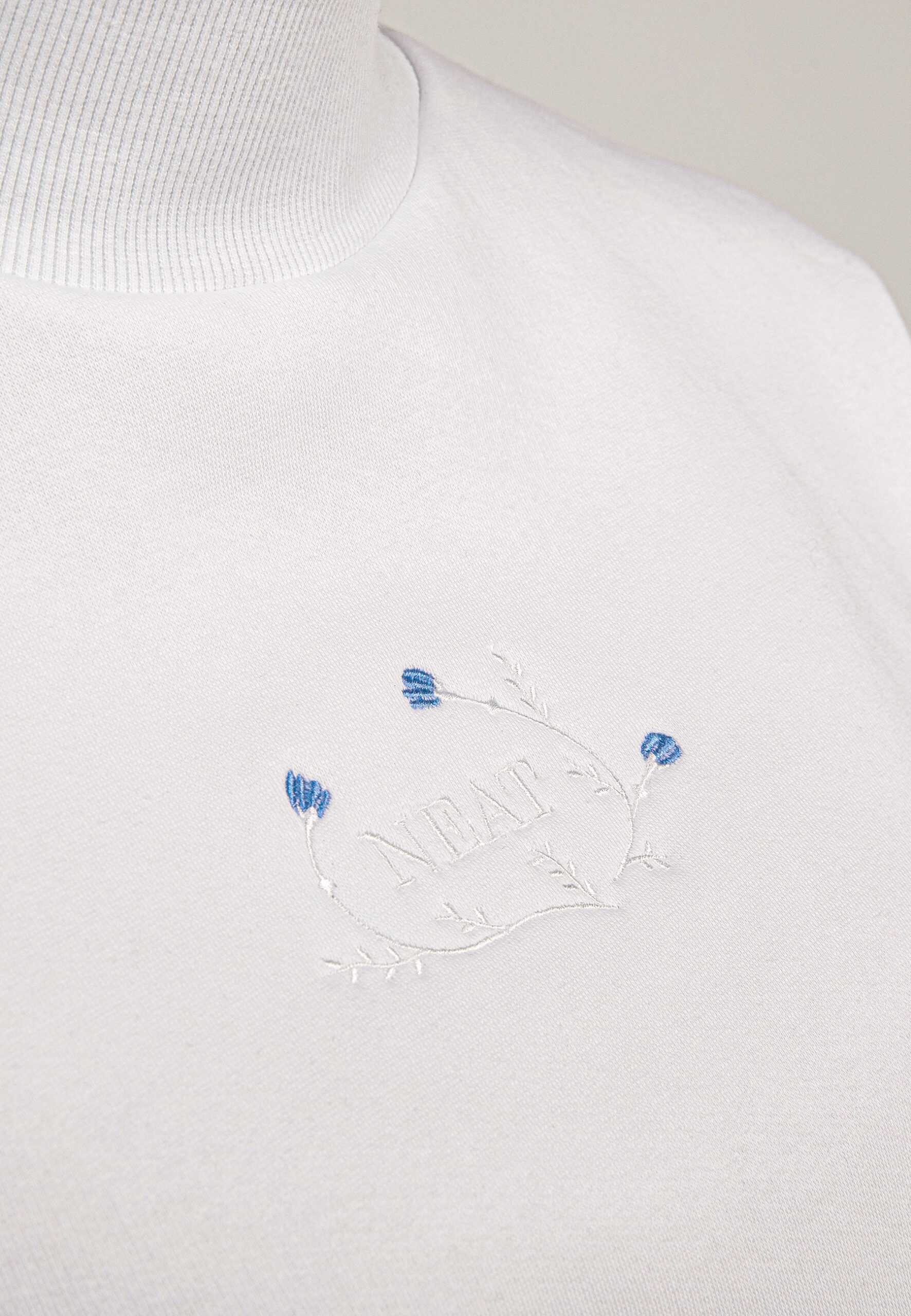 embroidery detail high-neck crop sweatshirt with a delicate blue floral embroidery and NEAT logo on the chest, paired with beige trousers.