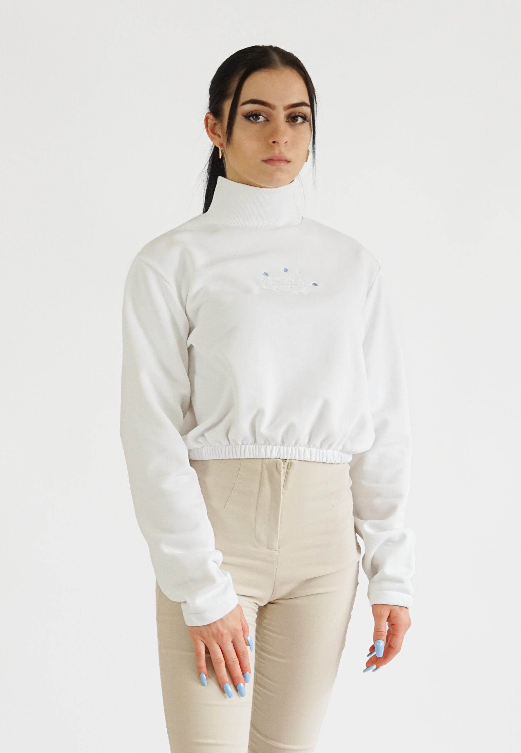 A model wearing a white high-neck crop sweatshirt with a delicate blue floral embroidery and NEAT logo on the chest, paired with beige trousers.
